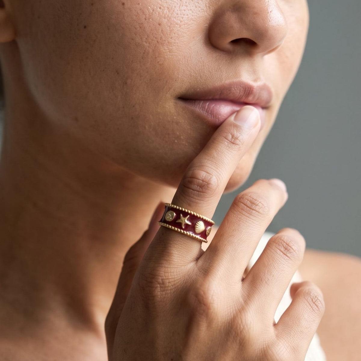 A close-up lifestyle shot of a woman wearing a wide band ring with red star details on a gold-toned band, paired with a cream-colored crochet knit top.